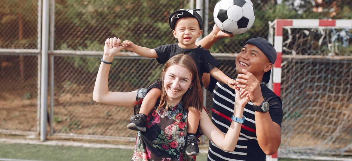 Positive smiling diverse parents giving piggyback ride to adorable toddler son while spending weekend together with football ball on sports ground on sunny day