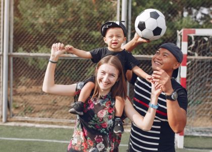 Positive smiling diverse parents giving piggyback ride to adorable toddler son while spending weekend together with football ball on sports ground on sunny day